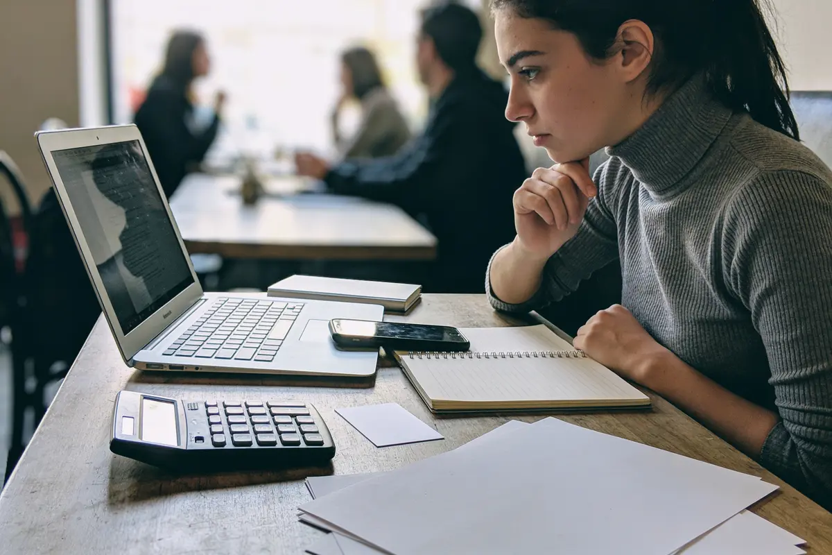 Jeune femme concentrée travaillant sur un ordinateur portable avec cahier, téléphone, calculatrice sur la table.