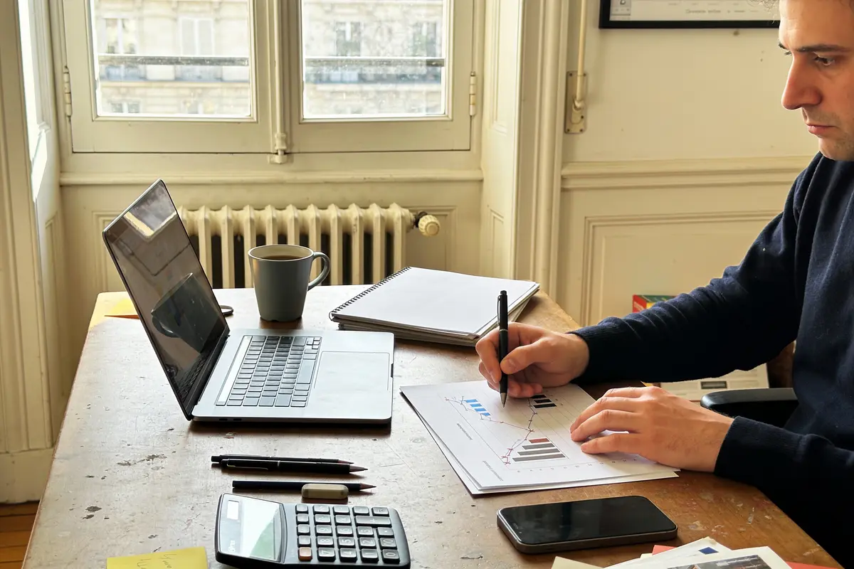 Homme travaillant sur un ordinateur portable avec graphiques, calculatrice et documents sur un bureau en bois.