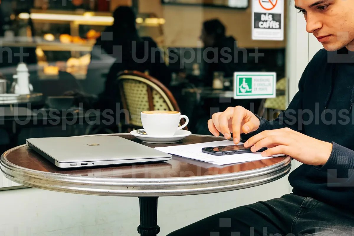 Homme utilisant un smartphone dans un café, avec un ordinateur portable et une tasse de café posés sur la table.