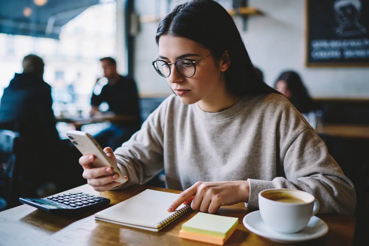 Femme avec lunettes utilise un smartphone et prend des notes dans un café, tasse de café à proximité.