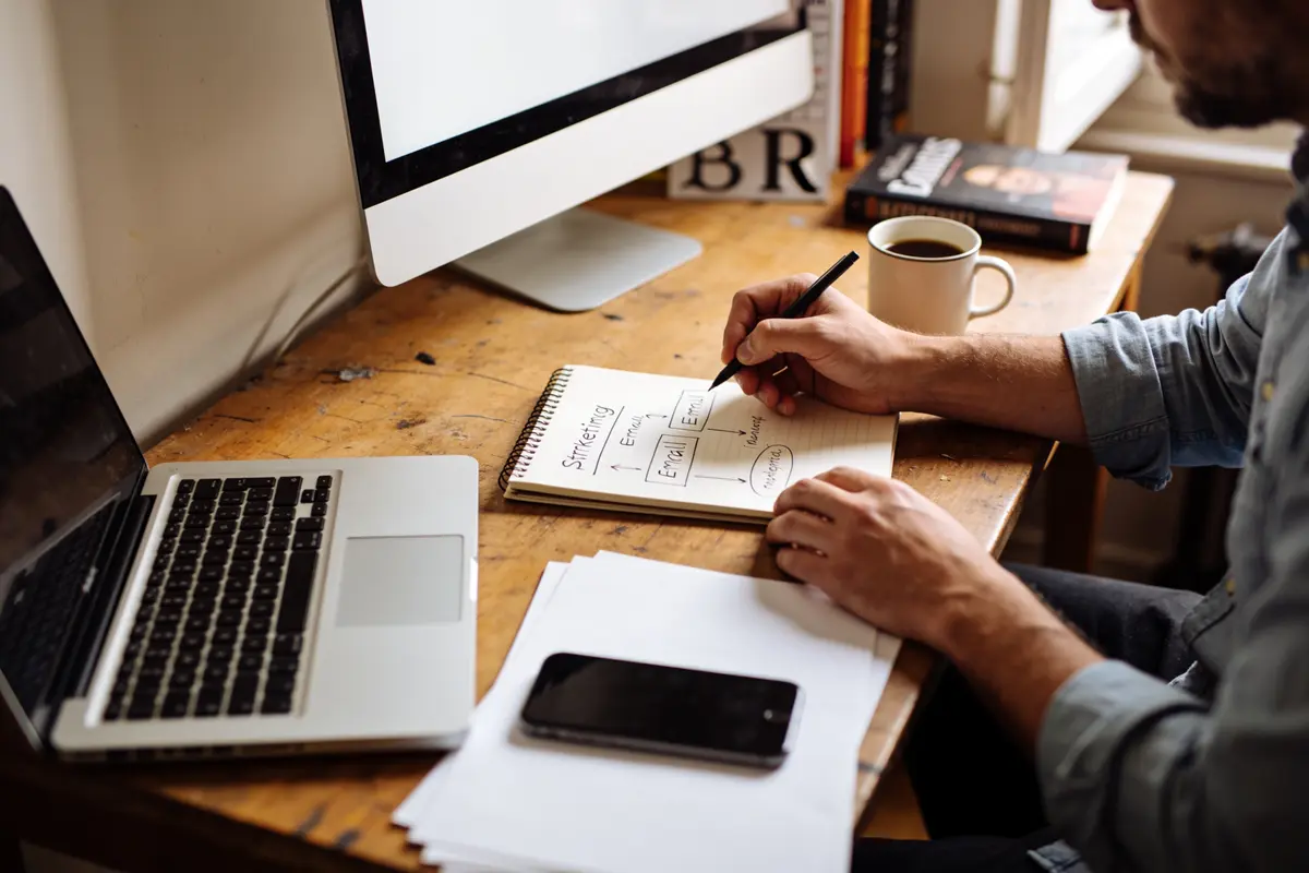 Personne écrivant des idées dans un carnet, avec un ordinateur portable, smartphone, et café sur un bureau.