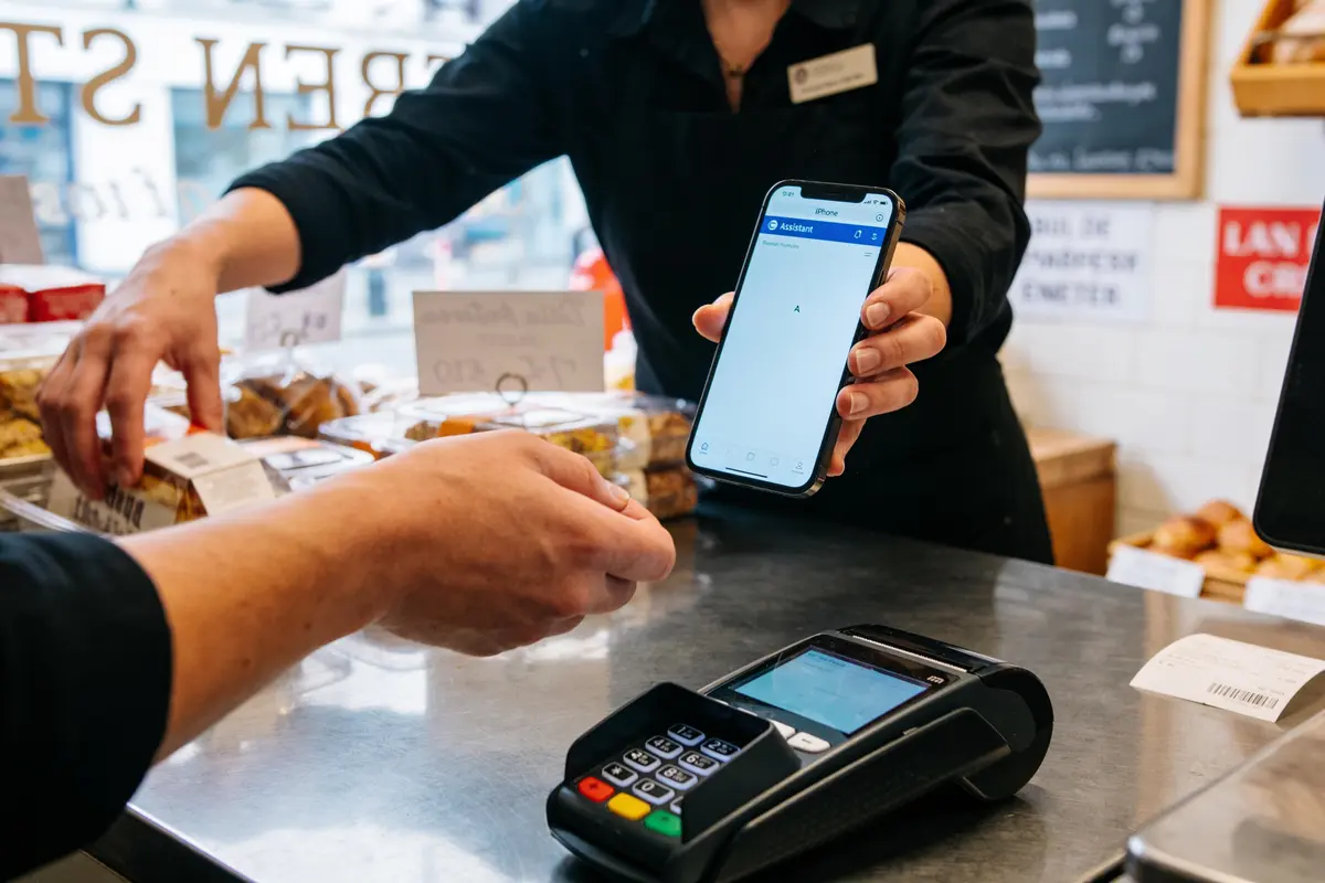 Personne payant via smartphone au comptoir d'une boutique, avec terminal de paiement électronique visible.
